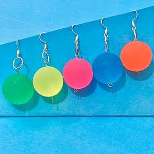 May include: Five pairs of colorful, round dangle earrings. The earrings are made of clear plastic and have a silver hook. The colors of the earrings are green, yellow, pink, blue, and orange.