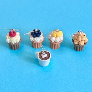 Op de afbeelding: Vijf paar hangende oorbellen met miniatuurvoedsel. De oorbellen zijn gemaakt van polymer clay en bevatten een cupcake met een rode kers erop, een cupcake met blauwe bessen erop, een cupcake met ananas erop, een cupcake met chocoladesprinkels erop en een witte koffiemok met een hartvormig ontwerp in het schuim.