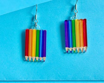 May include: A pair of colorful rainbow pencil earrings. Each earring features a rectangular design with a row of rainbow-colored pencils, with silver-colored fish hook ear wires. The earrings are set against a blue background.