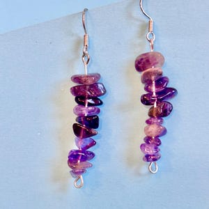 Può includere: Un paio di orecchini pendenti con perline di ametista in varie tonalità di viola. Le perline sono infilate su filo color argento, creando un effetto a cascata. Gli orecchini pendono da ganci per orecchie color argento.
