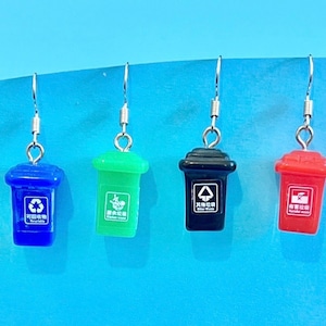 Könnte beinhalten: Vier Paar Ohrringe mit Miniatur-Mülleimern in verschiedenen Farben: blau, grün, schwarz und rot. Jeder Mülleimer hat ein anderes Recycling-Symbol und Text in Chinesisch.