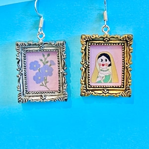 May include: A pair of silver and gold framed earrings with a floral and a cartoon portrait design. The floral design is on a pink background with purple flowers. The portrait design is on a pink background with a cartoon woman wearing a yellow dress.