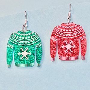 May include: A pair of dangle earrings featuring a green and red glitter sweater design with a white snowflake. The earrings have silver hooks.