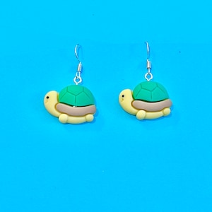 Puede incluir: Un par de pendientes de aro de plata con un dije de tortuga verde y amarilla. La tortuga está mirando hacia la derecha.