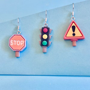 May include: Three pairs of dangle earrings featuring traffic signs. The earrings are silver with a stop sign, a traffic light, and a yellow triangle with an exclamation point.