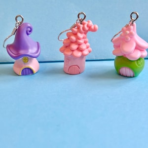 May include: Three whimsical house-shaped earrings. The earrings feature miniature houses with colorful details. One has a purple hat-like roof, another a pink tiled roof, and the third a pink roof with a green base. Each earring has a silver-tone hook.