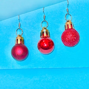 May include: Three red Christmas ornament earrings with gold accents. The ornaments are hanging from silver hooks.