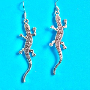 Puede incluir: Un par de pendientes de plata con forma de lagarto. Los lagartos están detallados con escamas texturizadas y tienen un diseño colgante.