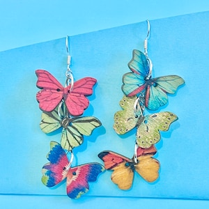 May include: A pair of dangle earrings featuring multiple layered butterfly charms. The butterflies are made of wood and have a variety of colourful patterns, including red, blue, green, and yellow.