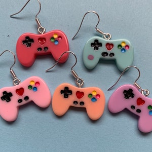 Puede incluir: Cinco pares de pendientes con forma de mando de videojuegos en colores pastel. Los colores son rojo, azul claro, rosa, melocotón y morado. Cada mando tiene botones negros, detalles coloridos y ganchos plateados.