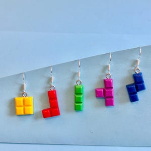 May include: Five earrings featuring Tetris block shapes in yellow, red, green, magenta, and blue. Each earring is suspended from a silver-coloured hook. The earrings are displayed on a light blue surface.