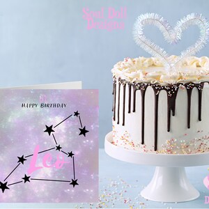 Puede incluir: Una tarjeta de cumplea&ntilde;os con tem&aacute;tica de galaxia rosa y morada con la constelaci&oacute;n de Leo y el texto "Feliz cumplea&ntilde;os". En el fondo hay un pastel blanco con glaseado de chocolate y chispas.