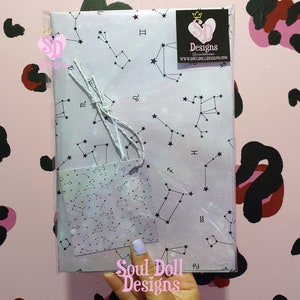 Puede incluir: Un papel de regalo blanco con un patr&oacute;n de constelaciones y una peque&ntilde;a etiqueta con un patr&oacute;n similar. El papel de regalo tiene un borde negro y el texto "Soul Doll Designs" est&aacute; impreso en la parte inferior. El papel de regalo est&aacute; envuelto alrededor de una caja de regalo.