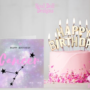 Puede incluir: Una tarjeta de cumplea&ntilde;os rosa con un fondo de galaxia p&uacute;rpura y blanco que presenta el signo del zod&iacute;aco C&aacute;ncer y el texto "Feliz cumplea&ntilde;os C&aacute;ncer". La tarjeta est&aacute; junto a un pastel rosa con velas doradas que deletrean "Feliz cumplea&ntilde;os".