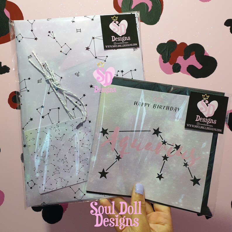 Puede incluir: Una tarjeta rosa y blanca con un dise&ntilde;o de constelaci&oacute;n y el texto "Happy Birthday Aquarius". La tarjeta est&aacute; envuelta en una funda de pl&aacute;stico transparente con una cuerda blanca atada. La tarjeta est&aacute; decorada con una peque&ntilde;a etiqueta con un dise&ntilde;o de constelaci&oacute;n. La tarjeta es de Soul Doll Designs.