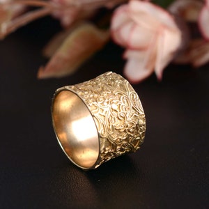 May include: A wide gold band ring with a detailed floral pattern. The ring is made of a smooth metal and has a polished finish.