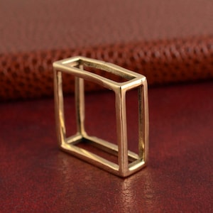 May include: A gold-colored, rectangular open-frame ring. The ring is made of a shiny metal and has a geometric design. The ring is displayed on a red surface.