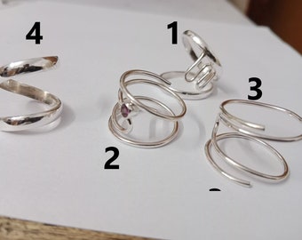 May include: Four silver rings, each with a unique design. The rings are numbered 1 through 4. Ring 1 is a simple, open design. Ring 2 has a spiral design with a small purple gemstone. Ring 3 is a double spiral design. Ring 4 is a simple, curved design.