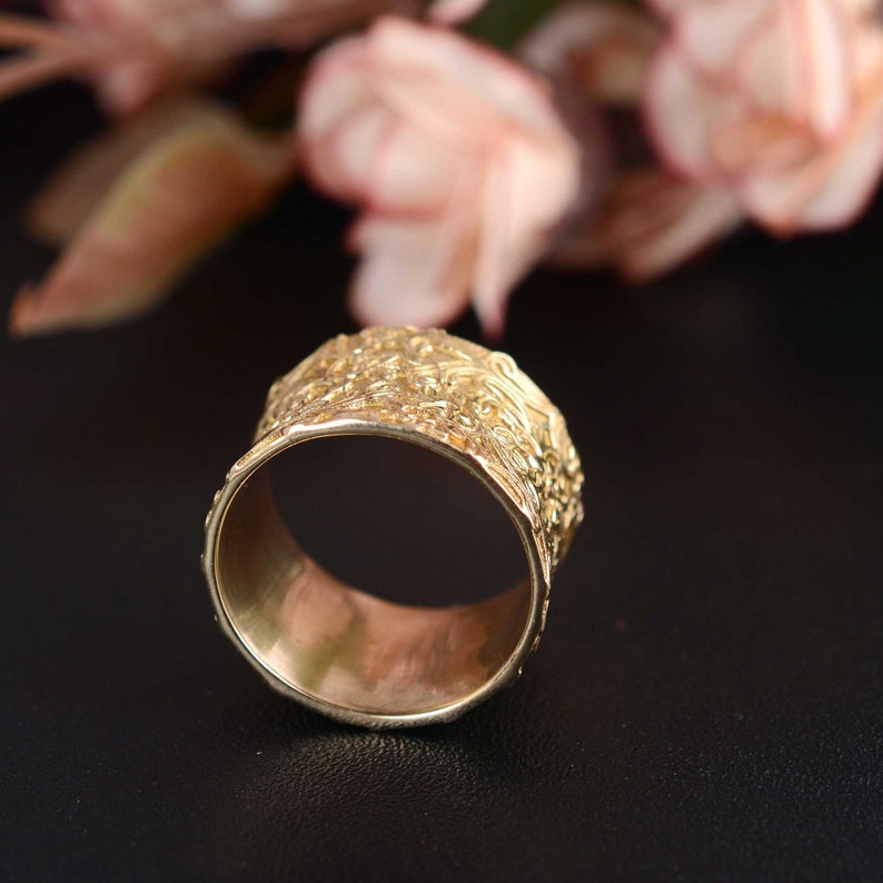 May include: A gold ring with a textured, ornate design. The ring is wide and has a smooth inner band.