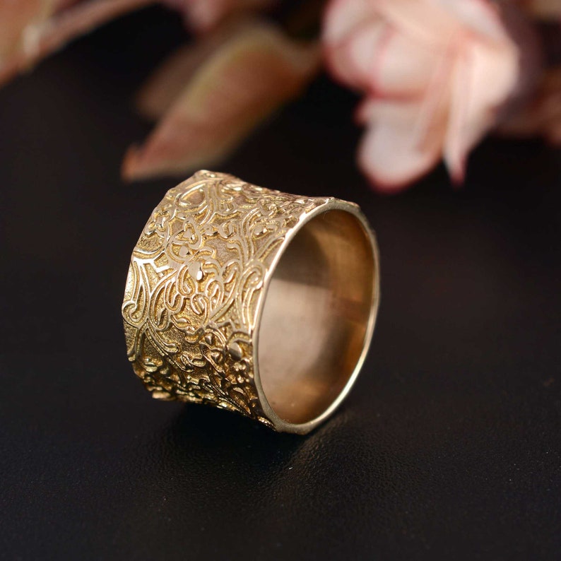 May include: A wide gold band ring with a detailed floral pattern. The ring is made of a smooth metal and has a polished finish.