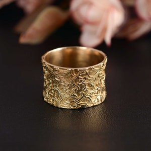 May include: A wide gold band ring with an intricate floral design. The ring is made of metal and has a smooth, polished finish.