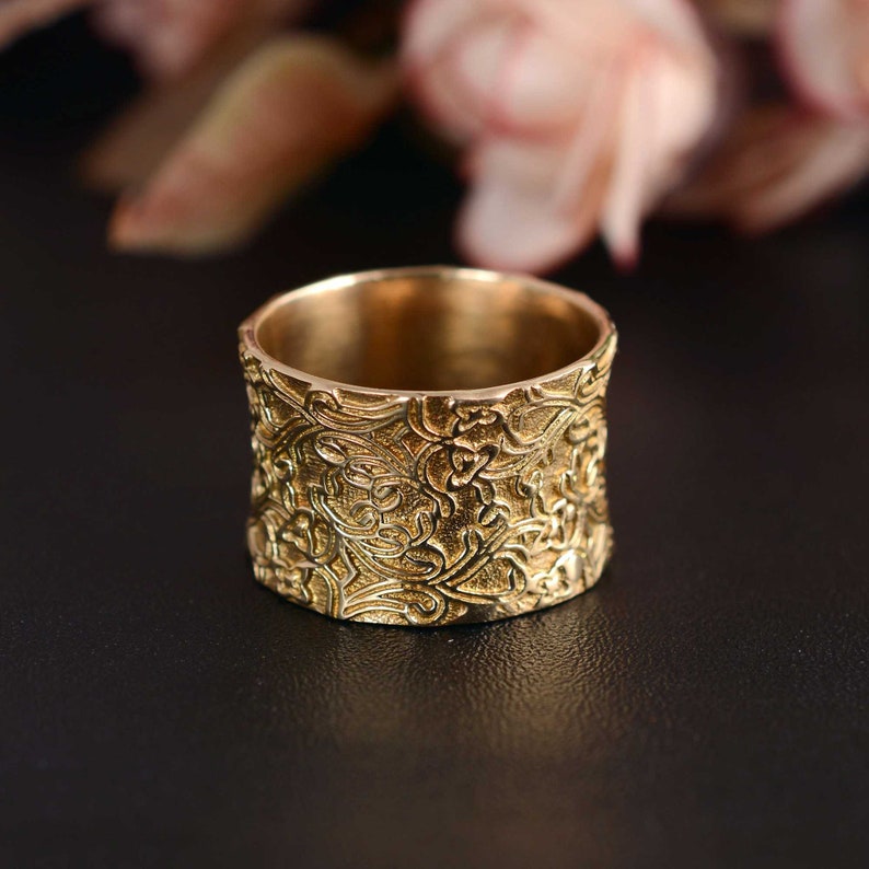 May include: A wide gold band ring with an intricate floral design. The ring is made of metal and has a smooth, polished finish.