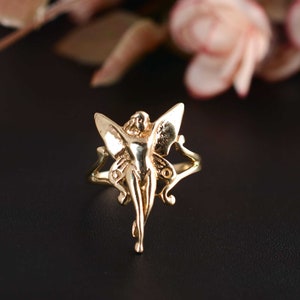 May include: A gold-colored ring featuring a fairy design. The fairy has outstretched wings and a slender body. The ring has intricate details and a polished finish. The background is dark, with a blurred floral element.
