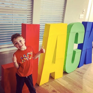 May include: A young boy wearing a red shirt with the word "JACK" in rainbow colors is standing in front of large cardboard letters that spell out "JACK". The letters are painted in different colors, including red, yellow, green, and blue.
