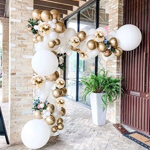 May include: A white and gold balloon arch with white and gold confetti balloons. The arch is decorated with greenery and flowers.