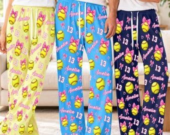 Könnte beinhalten: Drei Freizeithosen in Gelb, Blau und Marineblau, jeweils mit einem wiederkehrenden Muster aus gelben Softbällen mit rosa Schleifen, dem Namen Amelia in rosa Schreibschrift und der Zahl 13, alle mit weißem Kordelzug am Bund.