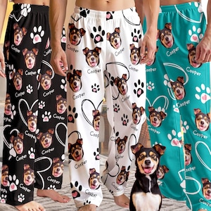 May include: Pajama pants in black, white, and teal feature a repeating pattern of a dog's face, paw prints, and the name "Cooper." The pants have a relaxed fit and are made of a soft, comfortable material. The pajama pants are suitable for lounging or sleeping.