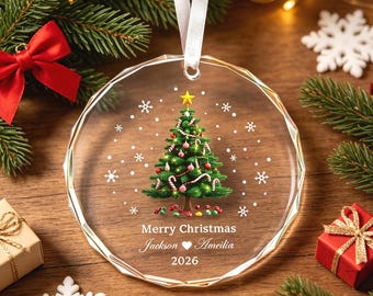 Pode incluir: Um enfeite de Natal circular de vidro transparente com bordas facetadas, apresentando uma árvore de Natal verde impressa com ornamentos, uma estrela amarela e flocos de neve brancos. O texto diz Merry Christmas, Jackson coração Amelia, 2026.