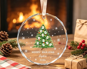 Pode incluir: Um ornamento de vidro transparente redondo e facetado pendurado por uma fita bege. O centro apresenta uma árvore de Natal verde decorada com peças de Mahjong, flocos de neve brancos e uma estrela dourada. O texto diz Merry Mahjong, Sophia, 2026.