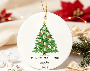 Pode incluir: Um ornamento redondo de cerâmica branca pendurado por um cordão dourado. O centro apresenta uma árvore de Natal verde decorada com peças de Mahjong e uma estrela amarela no topo. Abaixo da árvore, o texto diz MERRY MAHJONG, Sophia e 2026.