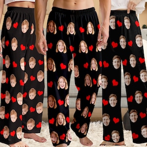 May include: Black pajama pants with a repeating pattern of red hearts and faces. The faces are of different people. The pajama pants are made of a soft material and have a drawstring waist.