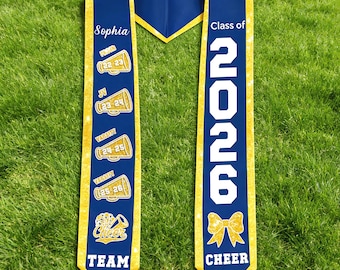 May include: A navy blue and gold graduation sash with the name "Sophia" and "Class of 2026" printed on it. The sash features cheerleading-themed graphics and the words "CHEER TEAM".