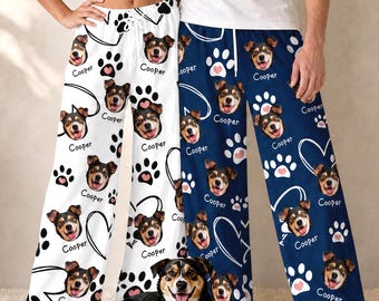 Pode incluir: Duas calças de pijama, uma branca e uma azul-marinho, com um padrão repetitivo de um rosto de cão, o nome Cooper, pegadas e contornos de corações. Um cão com marcas semelhantes está sentado em frente às calças no chão.