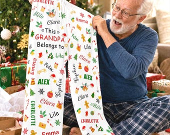 Pode incluir: Calças de pijama brancas com um padrão natalício repetitivo de ícones festivos coloridos e nomes como Charlotte, Milo, Clara, Emma, Felix, Lila, Alex e Sophia, com o texto This GRANDPA Belongs to impresso em vermelho e preto.