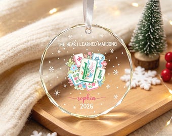 Pode incluir: Um ornamento de vidro circular e facetado pendurado por uma fita branca. O design apresenta um grupo central de peças de mahjong coloridas rodeadas por motivos florais, com o texto THE YEAR I LEARNED MAHJONG, o nome sophia e 2026.