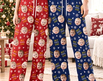 Pode incluir: Duas calças de pijama, uma vermelha e uma azul, com um padrão repetitivo de flocos de neve brancos e o rosto de um bebé. As calças vermelhas incluem o texto "BEST MAMA EVER" e as azuis "BEST PAPA EVER" ao lado de cada rosto.