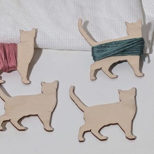 May include: Four wooden cat-shaped yarn holders. Two cats have pink and teal yarn wrapped around them. The other two cats are plain wood. The cats are on a white surface with a white cloth in the background.