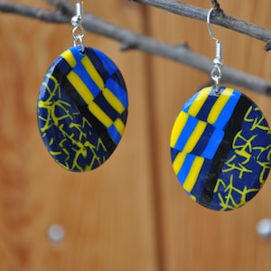 Peut inclure: Une paire de boucles d'oreilles ovales avec un motif géométrique bleu, jaune et noir. Les boucles d'oreilles sont en pâte polymère et ont des crochets argentés.