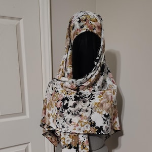 Puede incluir: Un hiyab con estampado floral con un velo facial negro. El hiyab presenta un fondo blanco con un patrón de flores rosas, marrones y negras. La bufanda está drapeada sobre un maniquí.