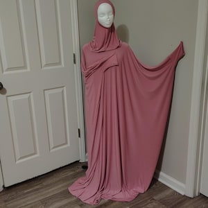 May include: A dusty rose-colored abaya, a modest garment, is displayed on a mannequin. The abaya features a matching head covering and a flowing, loose design. The fabric drapes elegantly, showcasing the garment's modest style and coverage.