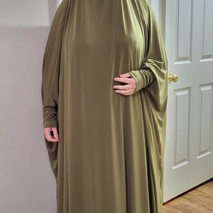 Puede incluir: Una prenda larga de tejido tipo jersey de color verde oliva con mangas largas. La prenda est&aacute; drapeada sobre el cuerpo de una persona, cubriendo su cabeza y brazos. El tejido es liso y tiene un ligero brillo.