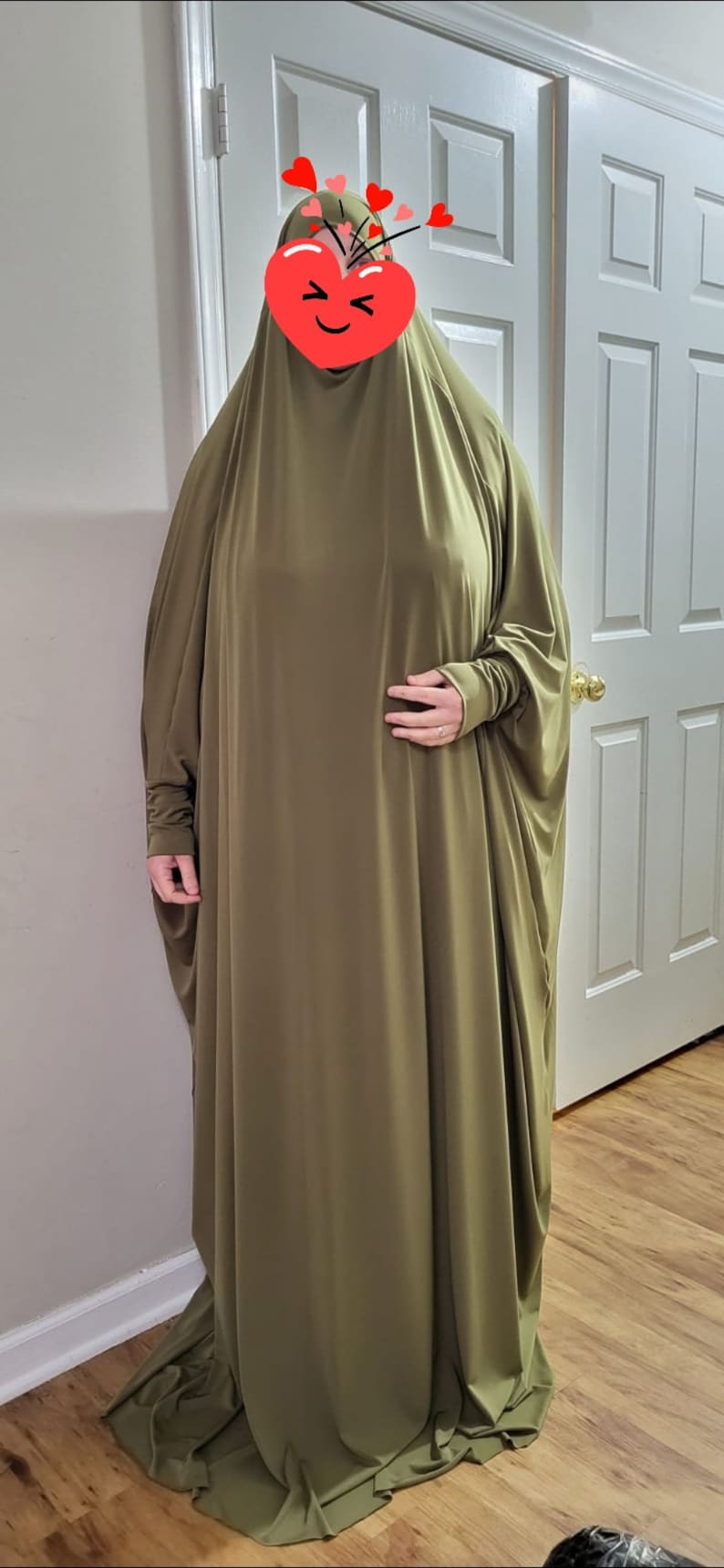 Puede incluir: Una prenda larga de tejido tipo jersey de color verde oliva con mangas largas. La prenda est&aacute; drapeada sobre el cuerpo de una persona, cubriendo su cabeza y brazos. El tejido es liso y tiene un ligero brillo.