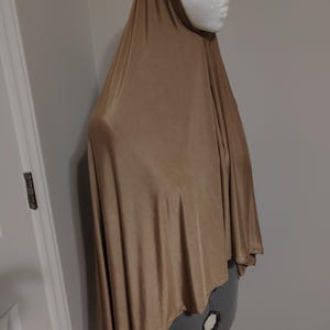 May include: A tan hijab, draped over a white mannequin head and a gray dress form. The fabric is smooth and flowing, with soft folds. The hijab is a solid color, and the overall style is modest and elegant.