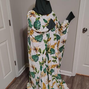 May include: A white dress with a tropical leaf pattern in green and yellow, displayed on a mannequin. The dress has long sleeves with dark green cuffs and a matching dark green head covering.