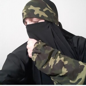 Puede incluir: Un niqab negro con una diadema de estampado de camuflaje. El niqab cubre la cara y el cuello, solo los ojos son visibles.
