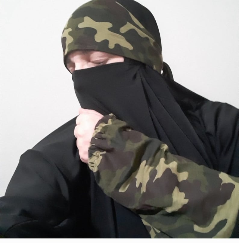 Puede incluir: Un niqab negro con una diadema de estampado de camuflaje. El niqab cubre la cara y el cuello, solo los ojos son visibles.